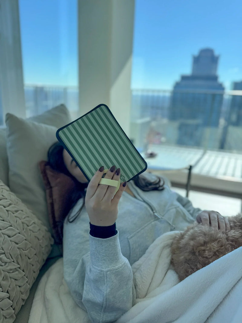 Person holding green striped tablet case on couch, city view in background, cozy setting