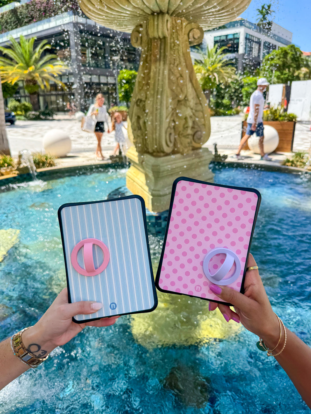 Two hands holding Bondi cases with pink and lavender grips in front of a fountain